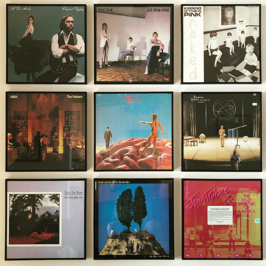 An arrangement of nine LP record covers on a wall, in which all the pictured figures are maintaining distance from one another. Artists include Al diMeola, The Jam, Kissing the Pink, Abba, Rush, Thomas Dolby, Tears for Fears, Elvis Costello & the Attractions, and John Trubee & the Ugly Janitors of America